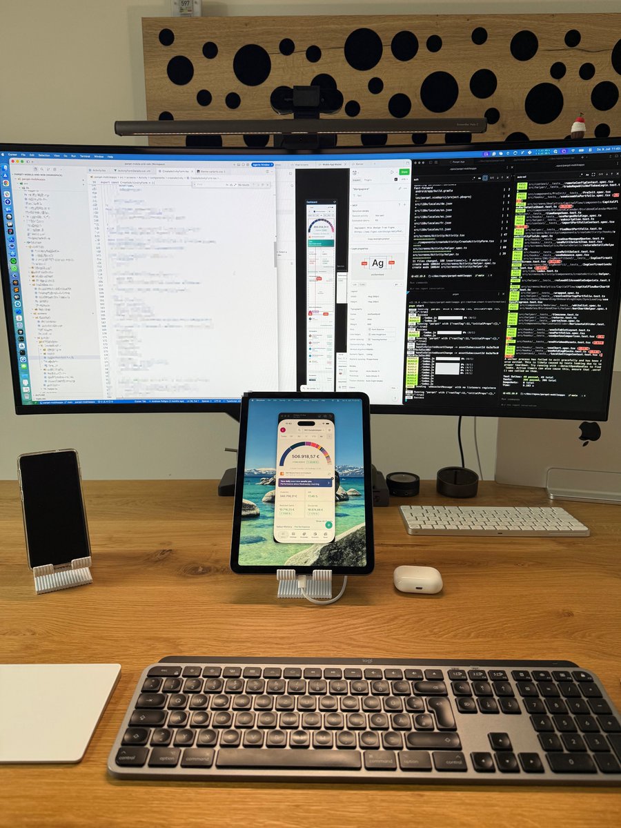 A developer desk with an ultrawide monitor and an iPad in portrait on a stand serving as a second display, showing a mobile app preview